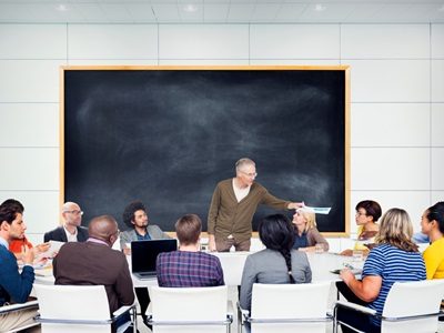A gray haired man stands in front of a large blackboard and addresses a multiethnic group of students.  The students are sitting around a large table.